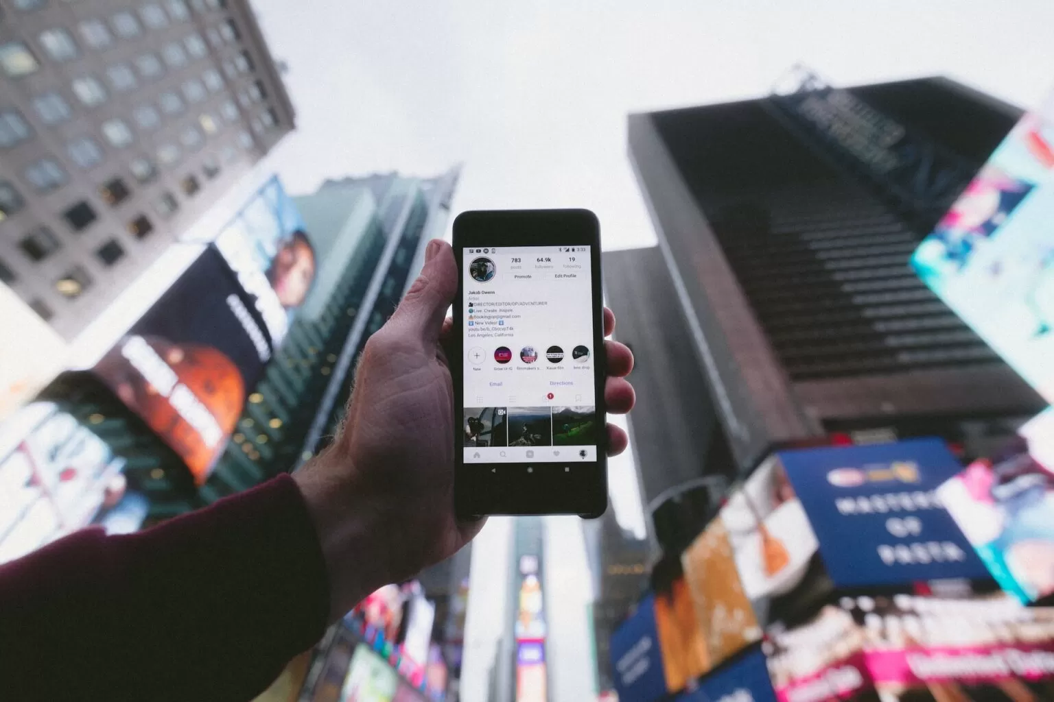 "Mão segurando o telemóvel com perfil de empresa aberto no meio da cidade de Nova Iorque. Agência de marketing digital especializada em publicidade em dispositivos móveis e campanhas publicitárias para empresas locais." Palavras-chave: marketing digital, publicidade em dispositivos móveis, campanhas publicitárias, empresa de marketing, Nova Iorque, perfil de empresa.