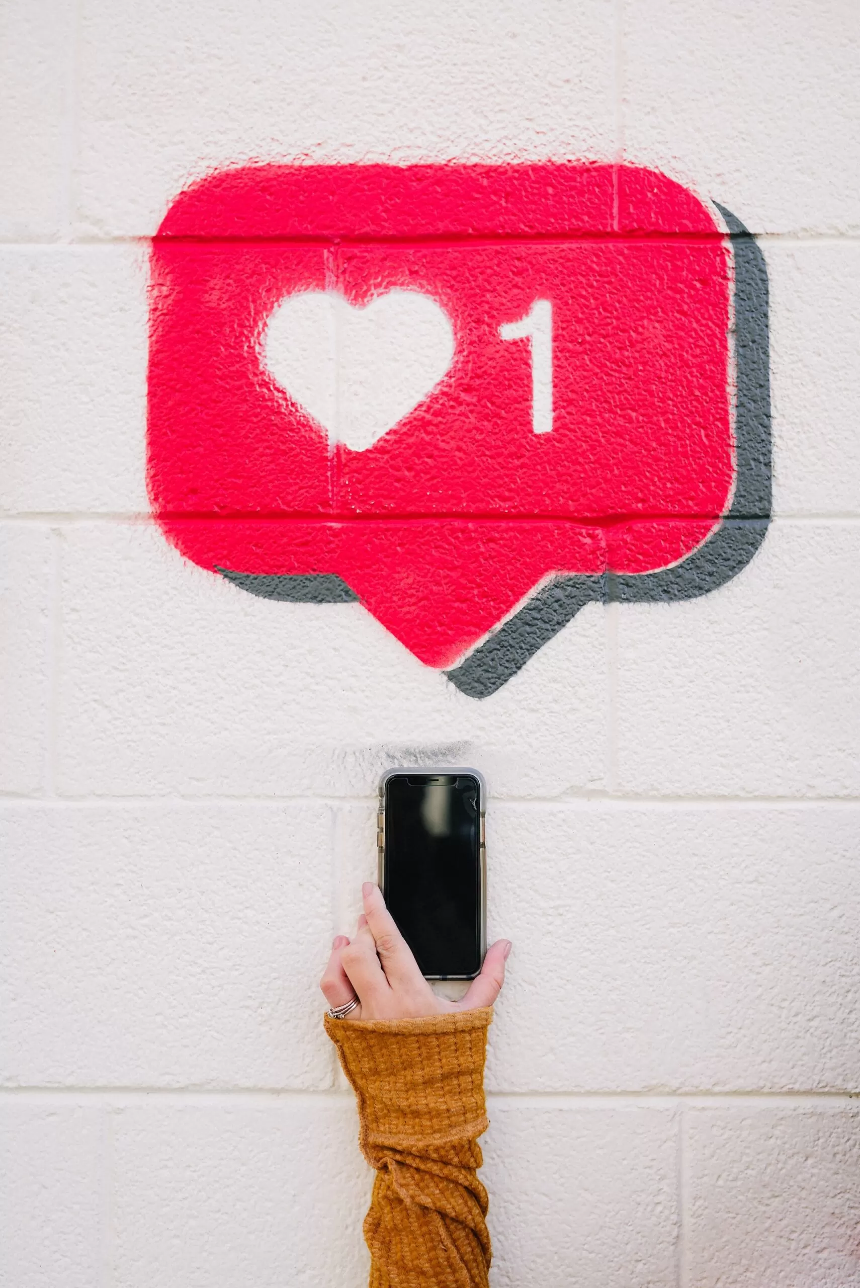 Mão segurando telemóvel com grafitti de coração (like) ao fundo - Serviços de Gestão de Redes Sociais